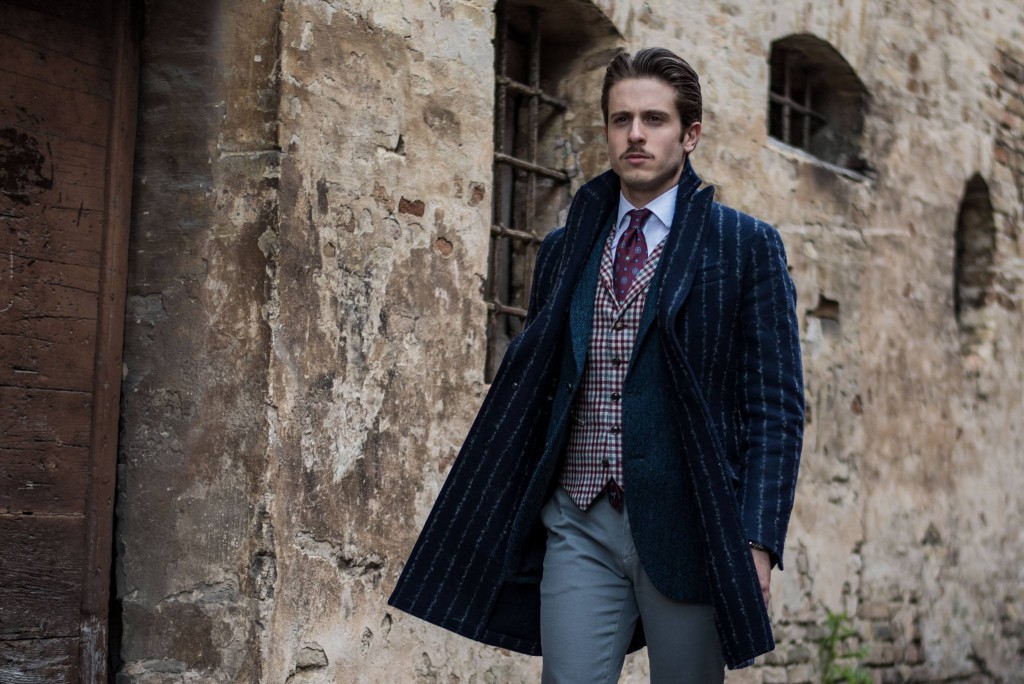 The ice colored trousers by Yan Simmon match the gray and burgundy checkered waist coat, presented here with a blue herringbone jacket. The blue coat by Hevò is made of wool and pinstriped fabric