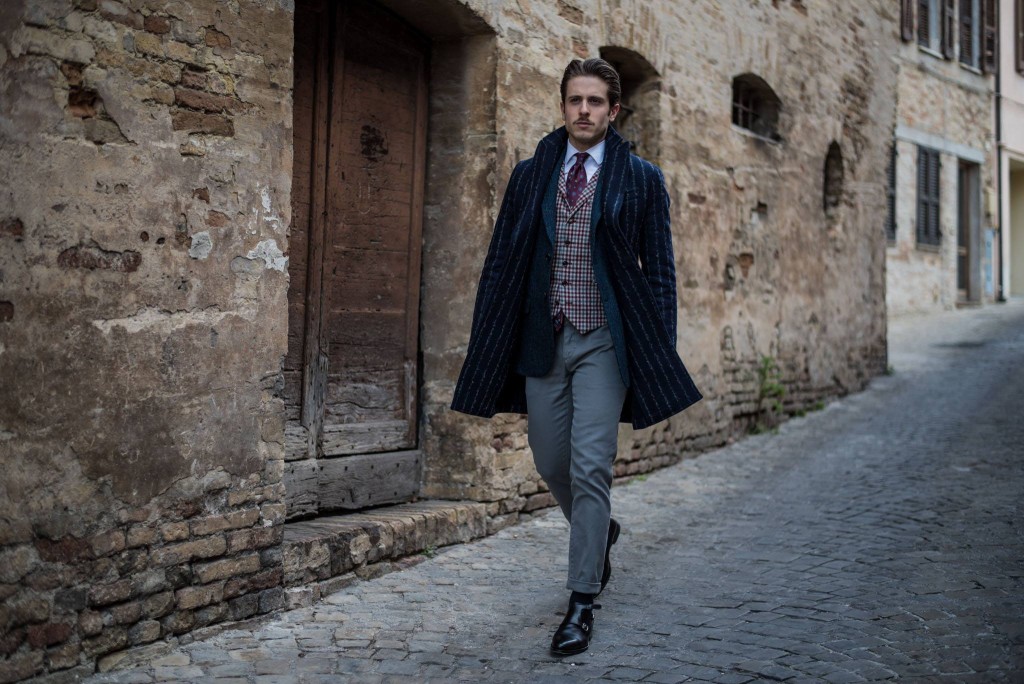 The ice colored trousers by Yan Simmon match the gray and burgundy checkered waist coat, presented here with a blue herringbone jacket. The blue coat by Hevò is made of wool and pinstriped fabric